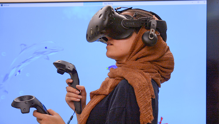 ​  A female student is wearing an HTC Vive VR headset and holding VR remote controls while standing in front of a TV screen depicting an underwater scene in a VR application. 