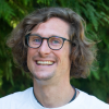 bespectacled man; exuberant smile, goatee; green leaves frame wild hair